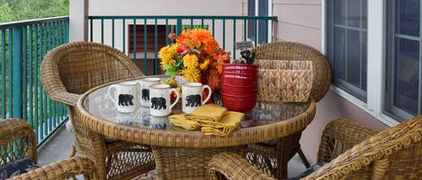 A wicker table with four mugs, a vase of flowers, and a red bucket on a balcony.