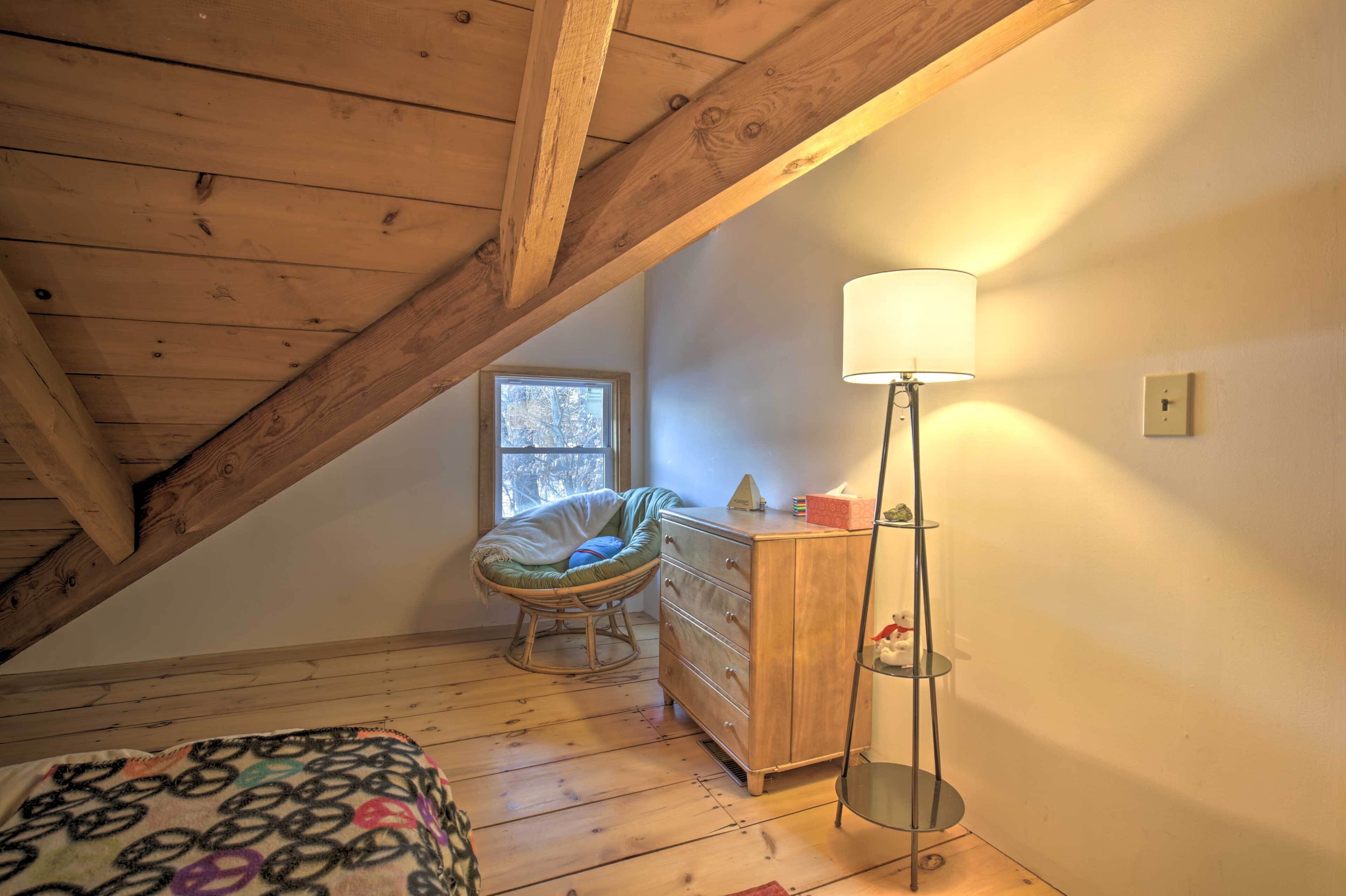Ein Zimmer mit schrägen Wänden, Holzbalken, einem Korbstuhl, einem Schminktisch und einer Stehlampe.