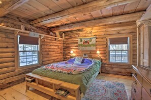 A log cabin bedroom with a bed, two bedside lamps, a window, a bench, and a rug. - Cabin: Private Hot Tub, Walk to Pats Peak Ski Area (Henniker)