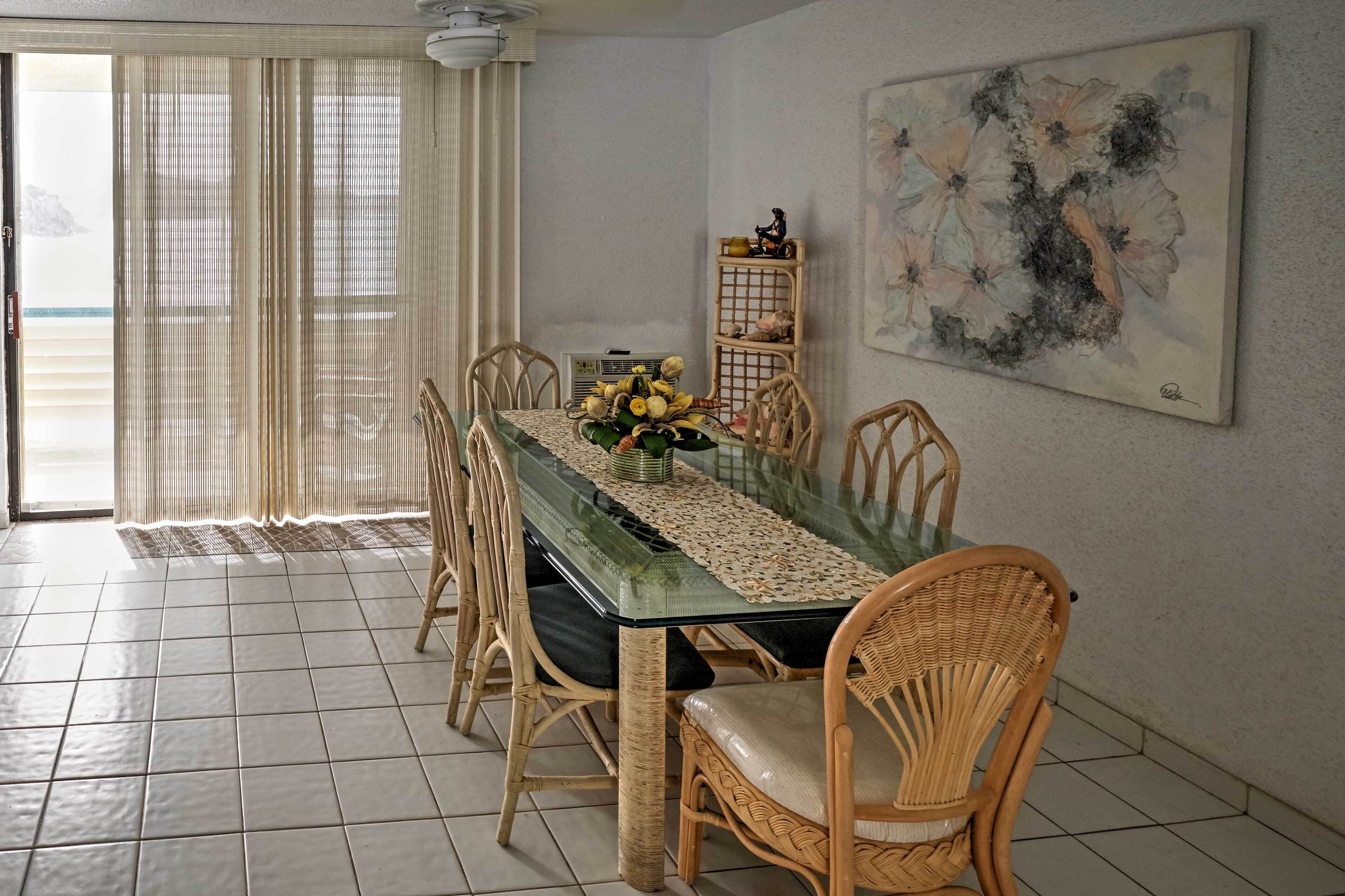 Un comedor con una mesa de vidrio, sillas de mimbre, un mantel de encaje y un cuadro floral en la pared.