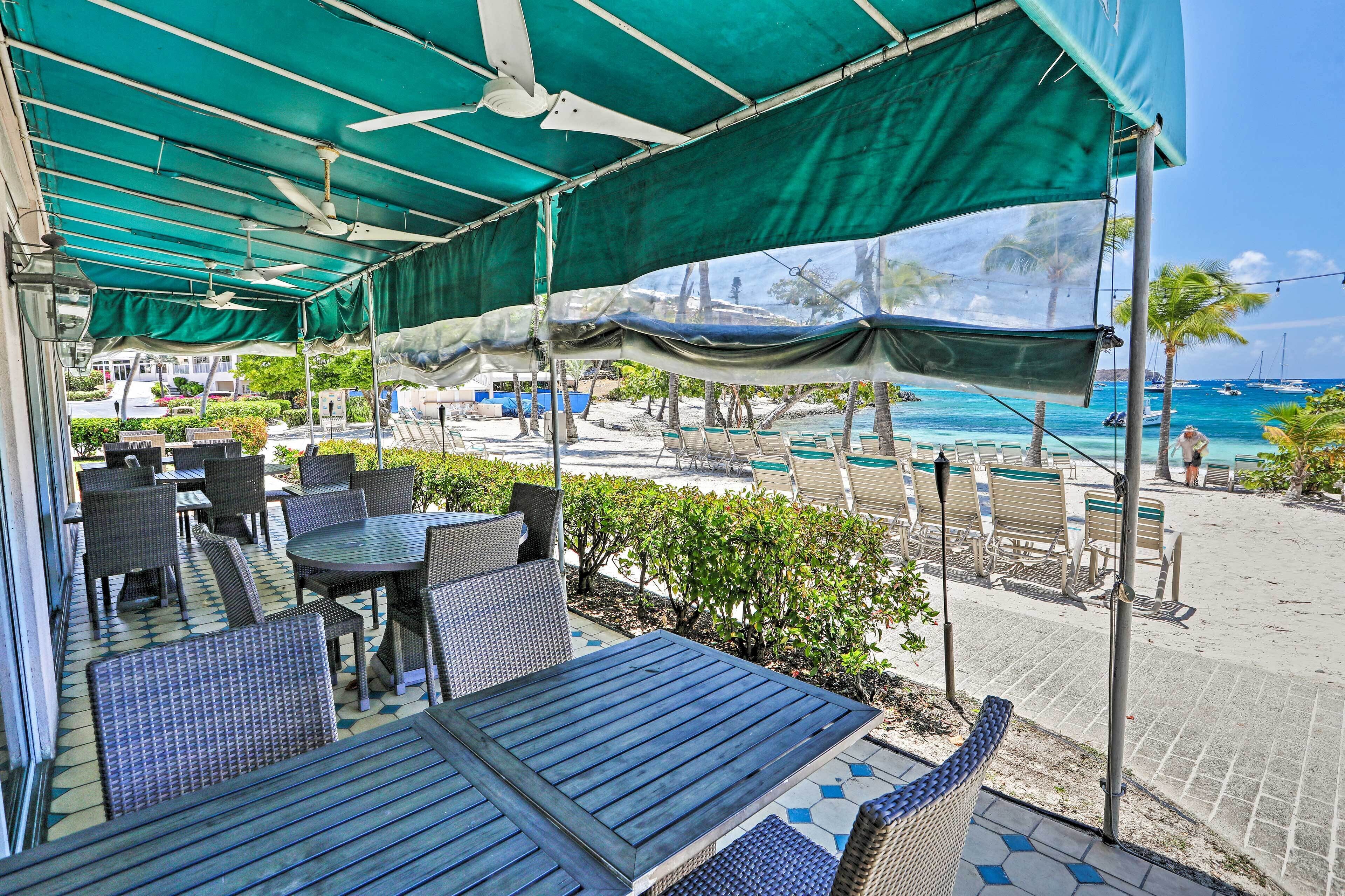 Un espace extérieur aménagé avec des tables et des chaises, abrité par une pergola verdoyante, et offrant une vue sur une plage bordée de palmiers et de bateaux.
