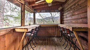 Innenansicht einer Holzhütte mit Esstisch und Stühlen, einem Deckenventilator und einem großen Fenster mit Blick auf Bäume und Berge.