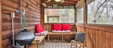 Una baita in legno con una ventola a soffitto, una finestra vista alberi, un barbecue e una panca con cuscini rossi.
