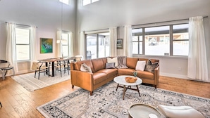 A spacious living room with a leather sectional sofa, a round coffee table, and a patterned rug. There are multiple windows providing natural light, and a dining area with a table and chairs is visible in the background.