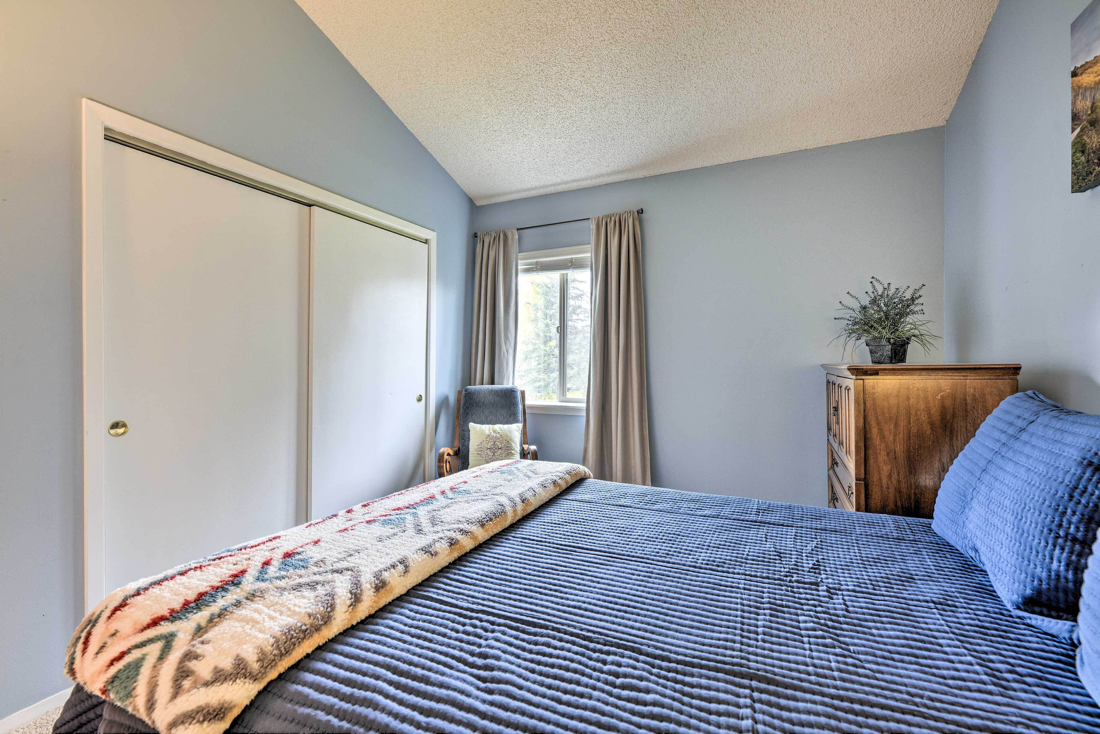 A bedroom with a bed, a chair, a wooden dresser, a window with curtains, and a door.