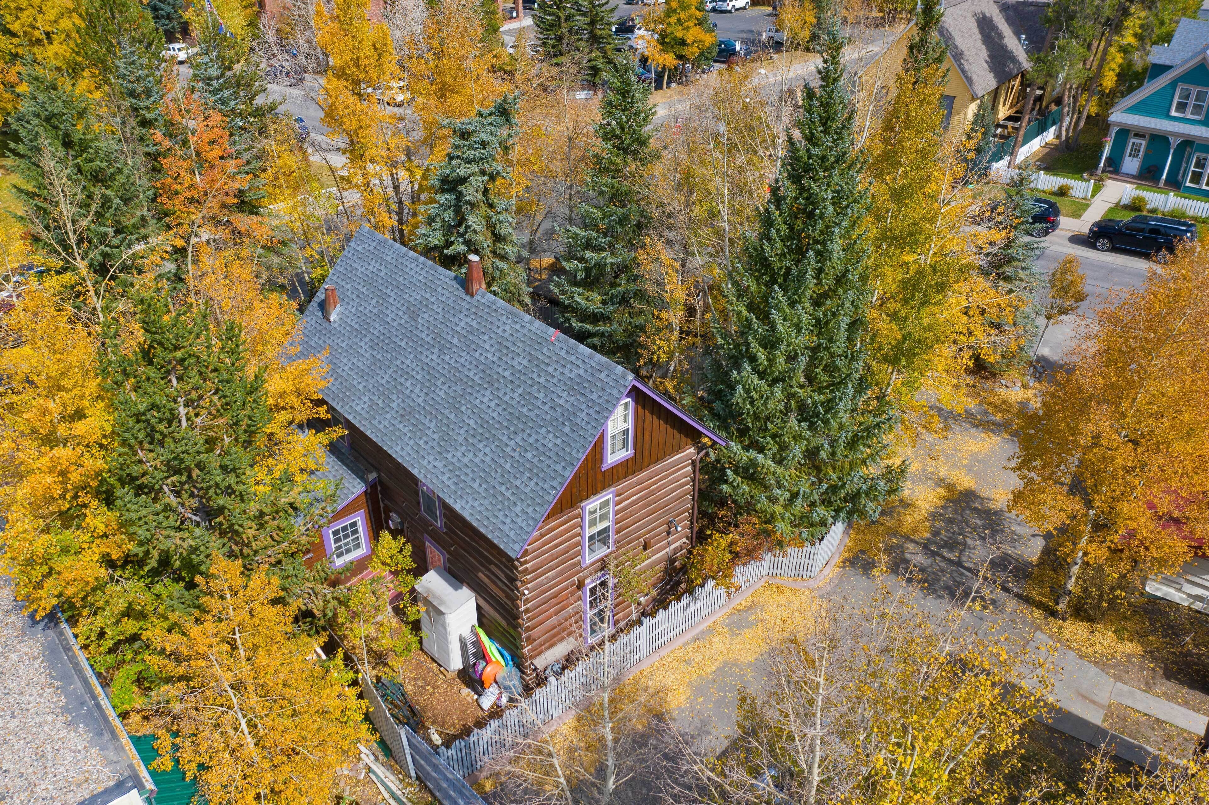A log cabin with a sloped roof, surrounded by autumn foliage, and a white picket fence.