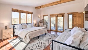 Un dormitorio con una cama grande, otra más pequeña, una mesita de noche, una lámpara, un sillón de fibras naturales y vistas al exterior a través de amplias ventanas.
