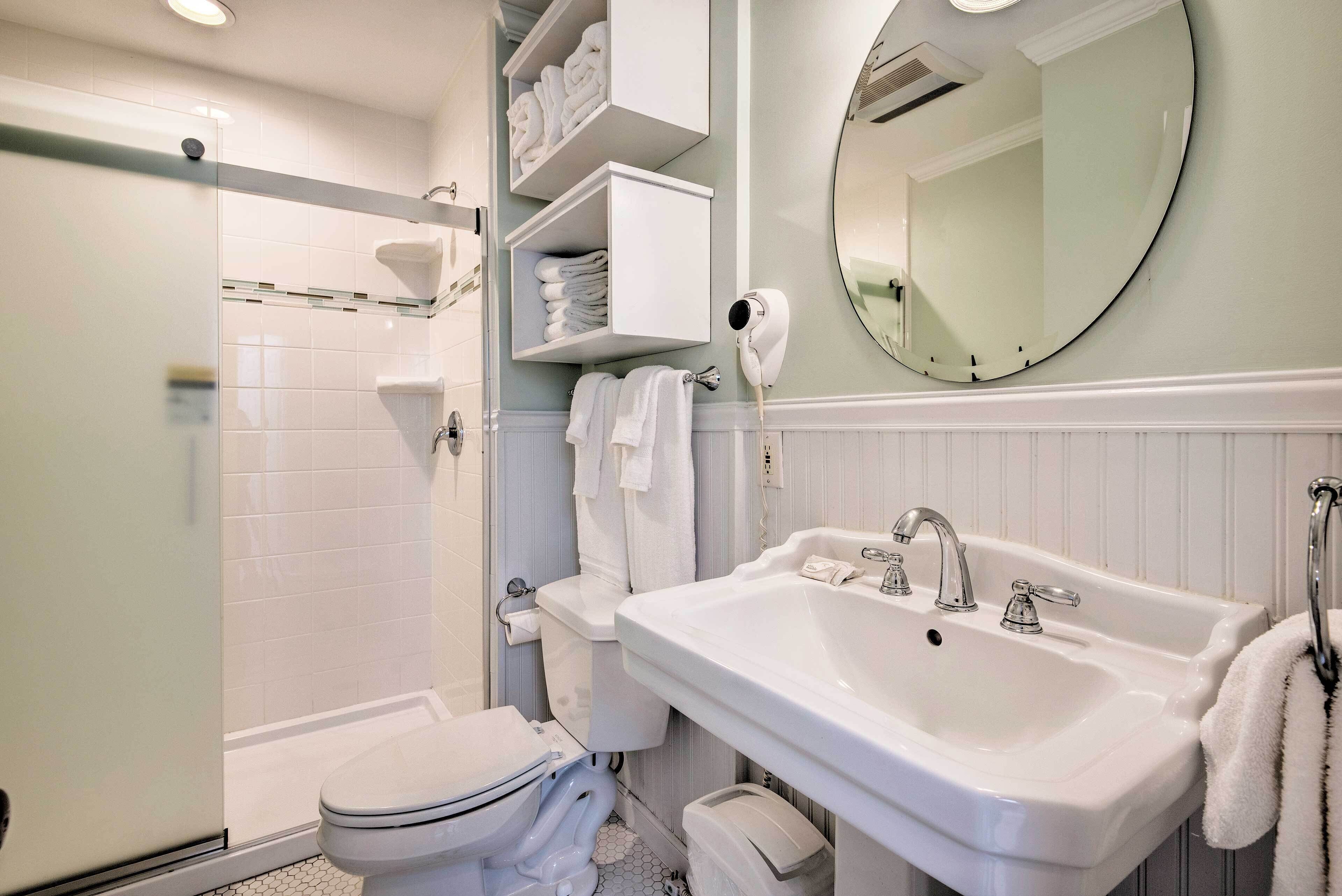 Un baño con inodoro, lavamanos y ducha con mampara de vidrio. Hay repisas con toallas y un secador de pelo fijado en la pared.