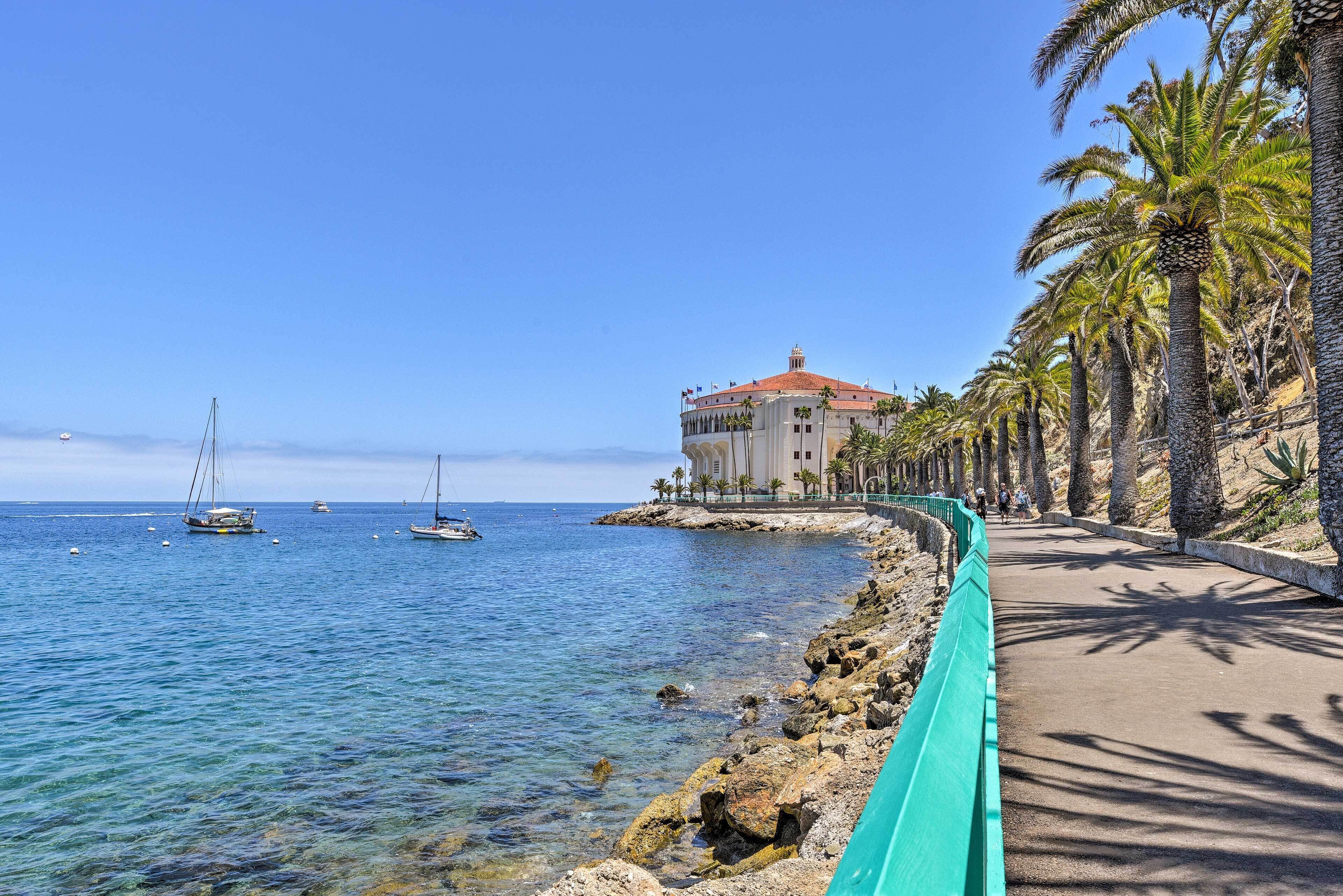 A coastal road with a building on the right, clear blue water, and boats in the distance.