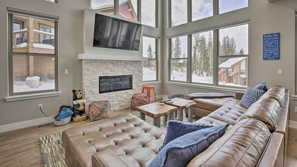 A spacious living room with a large window, a fireplace, a leather sofa, and a television mounted on the wall. - Winter Park Area Retreat, Walk to Ski Shuttle (Fraser)