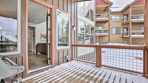 A wooden deck with a metal railing, a glass door leading inside, and a view of a multi-story building with balconies. - Winter Park Area Retreat, Walk to Ski Shuttle (Fraser)