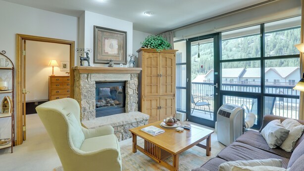 Una sala de estar con chimenea, un aparador de madera, un sofá, una mesa y un ventanal con vistas a casas y árboles.