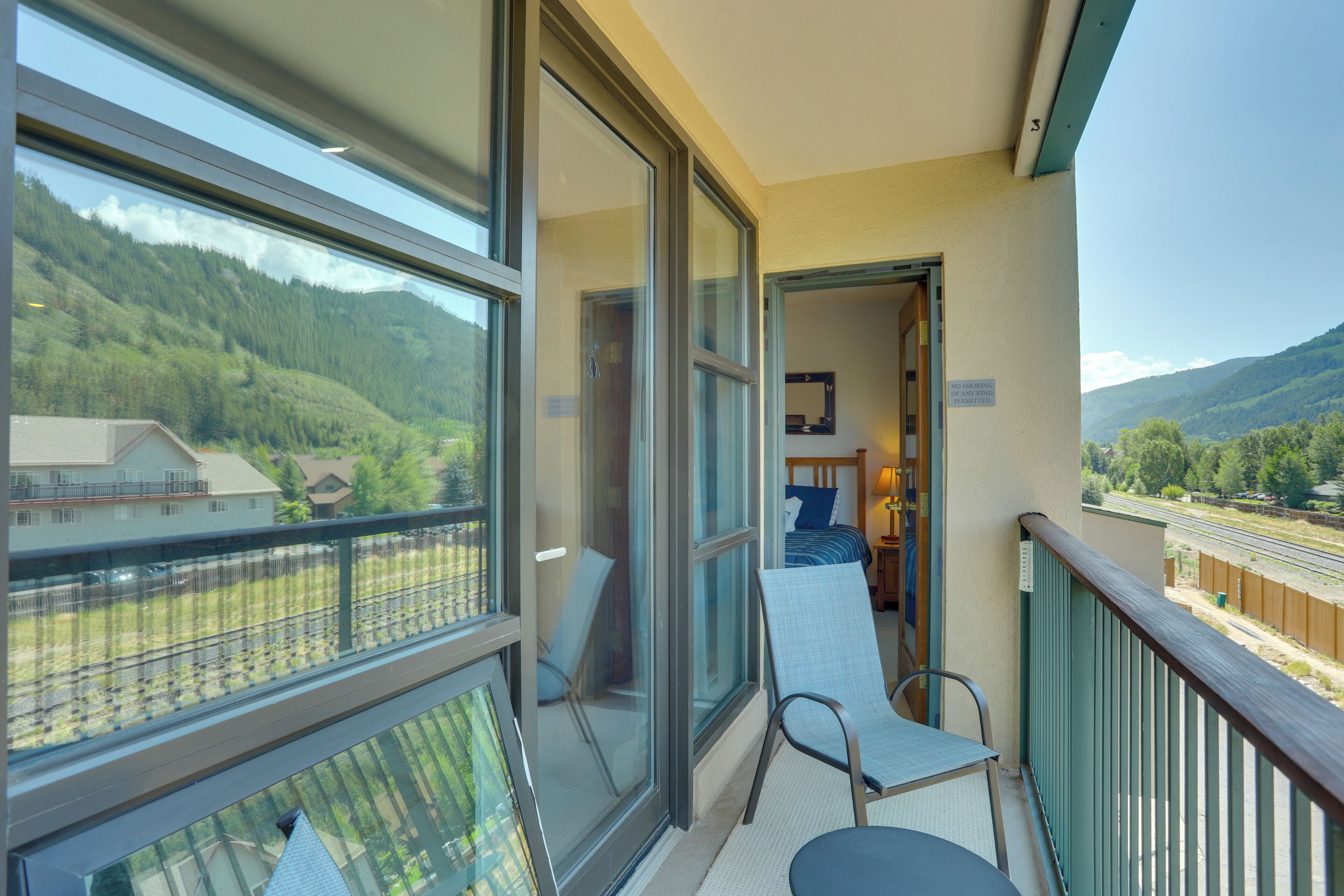 A balcony with a table and chairs, a bed with a blue pillow, and a view of a mountainous landscape.