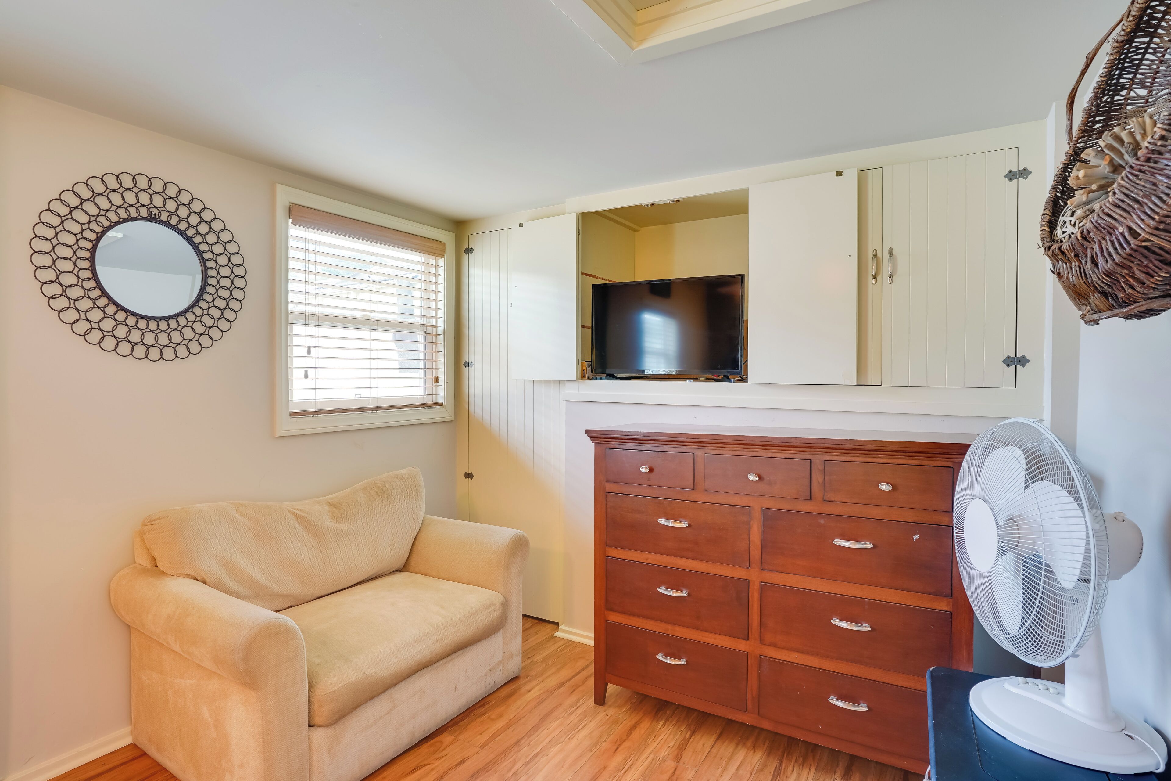 A room with a beige sofa, a wooden dresser, a TV, and a fan.