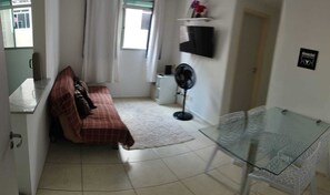 Ein kleiner, gut beleuchteter Raum mit einem Bett, einem Glastisch, einem Standventilator und einem Fernseher an der Wand.