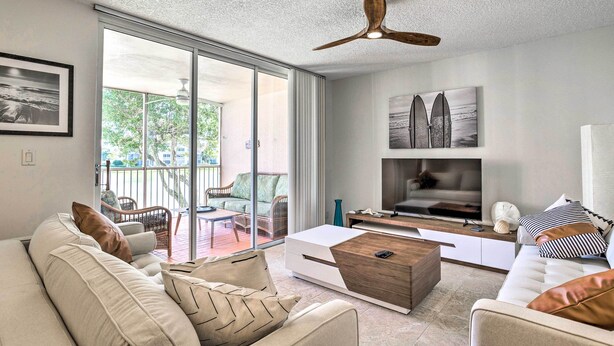 A modern living room with a sofa, coffee table, and TV. There is a balcony with chairs and a view of greenery. - Jupiter Bay Condo w/ Pool < Half Mile to Beach! (Jupiter)