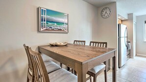 A dining area with a wooden table and chairs, a clock on the wall, and a kitchen in the background. - Jupiter Bay Condo w/ Pool < Half Mile to Beach! (Jupiter)