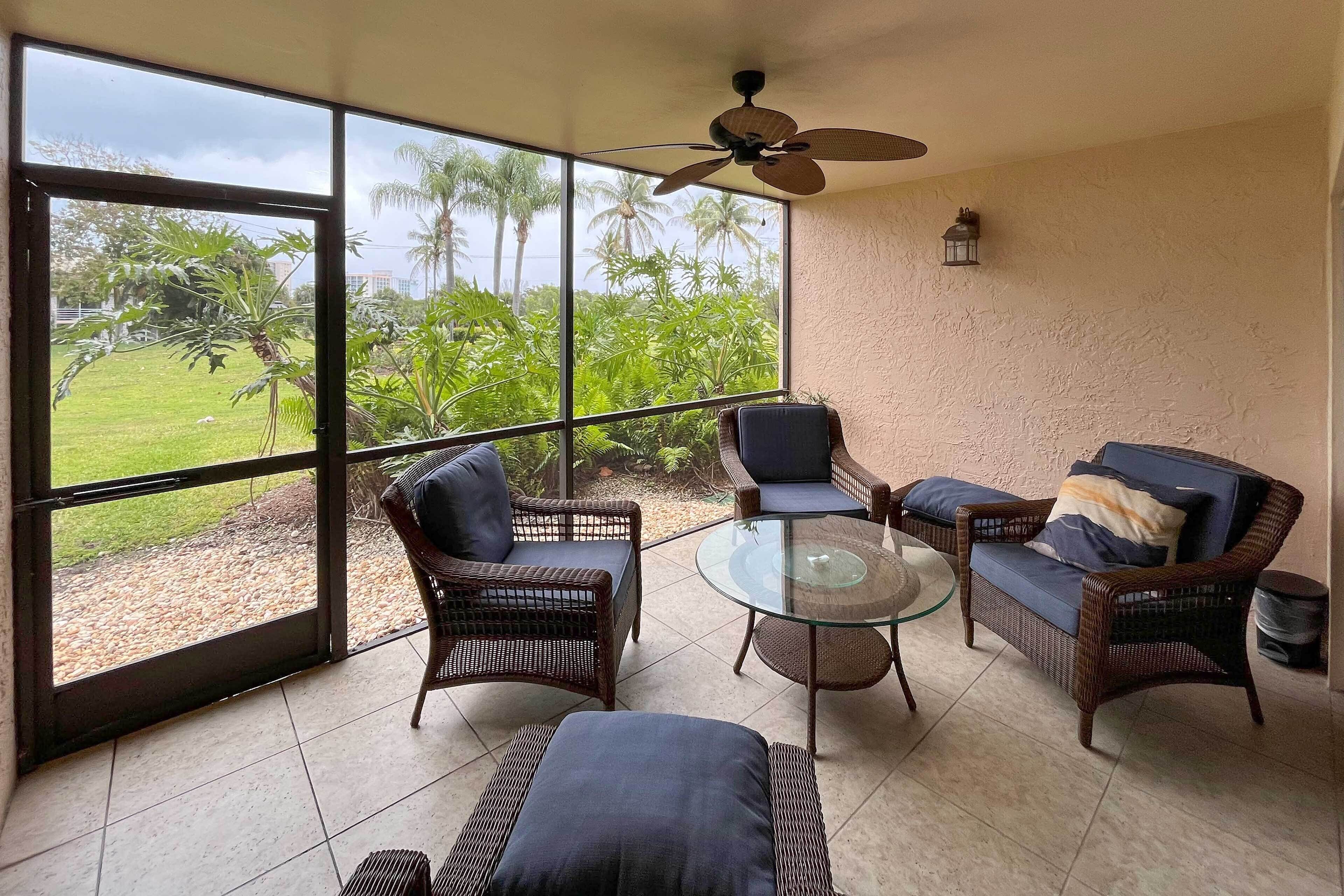 A room with wicker furniture, a glass table, and a view of a grassy area with palm trees.