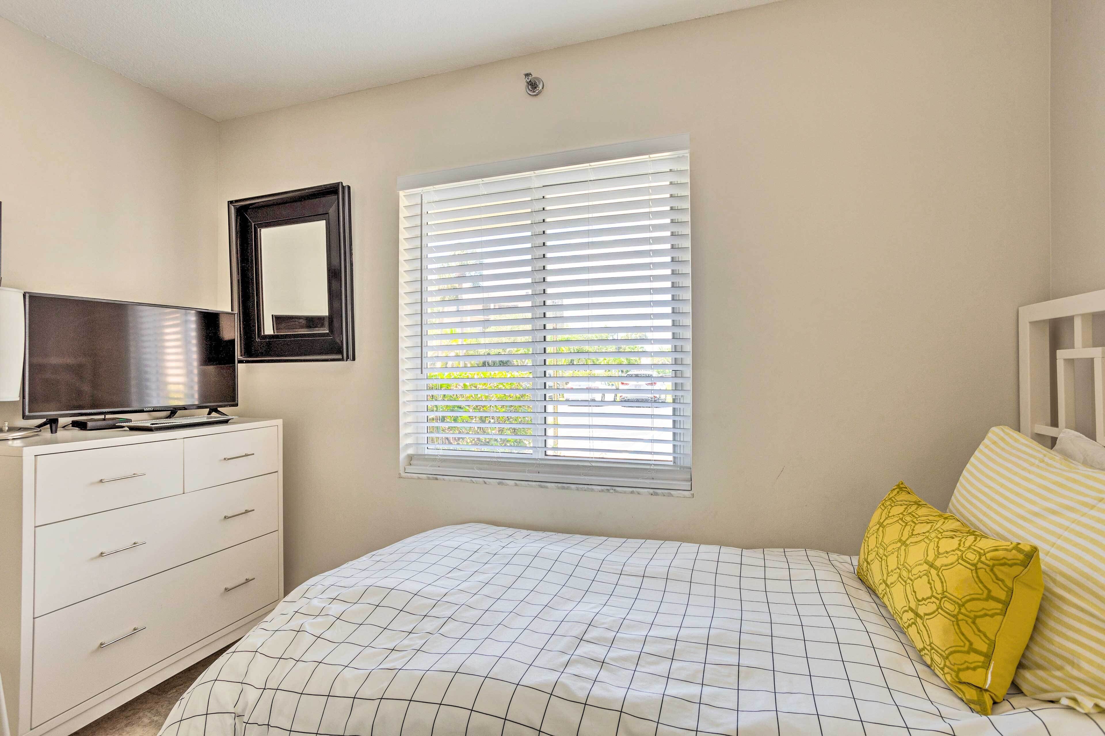 A bedroom with a bed, a TV on a dresser, a window with blinds, and a wall-mounted mirror.