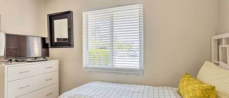 A bedroom with a bed, a TV on a dresser, a window with blinds, and a wall-mounted mirror.