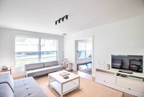 A modern living room with a grey sofa, a white coffee table, a flat-screen TV on a white stand, and large windows offering a view of the outdoors.