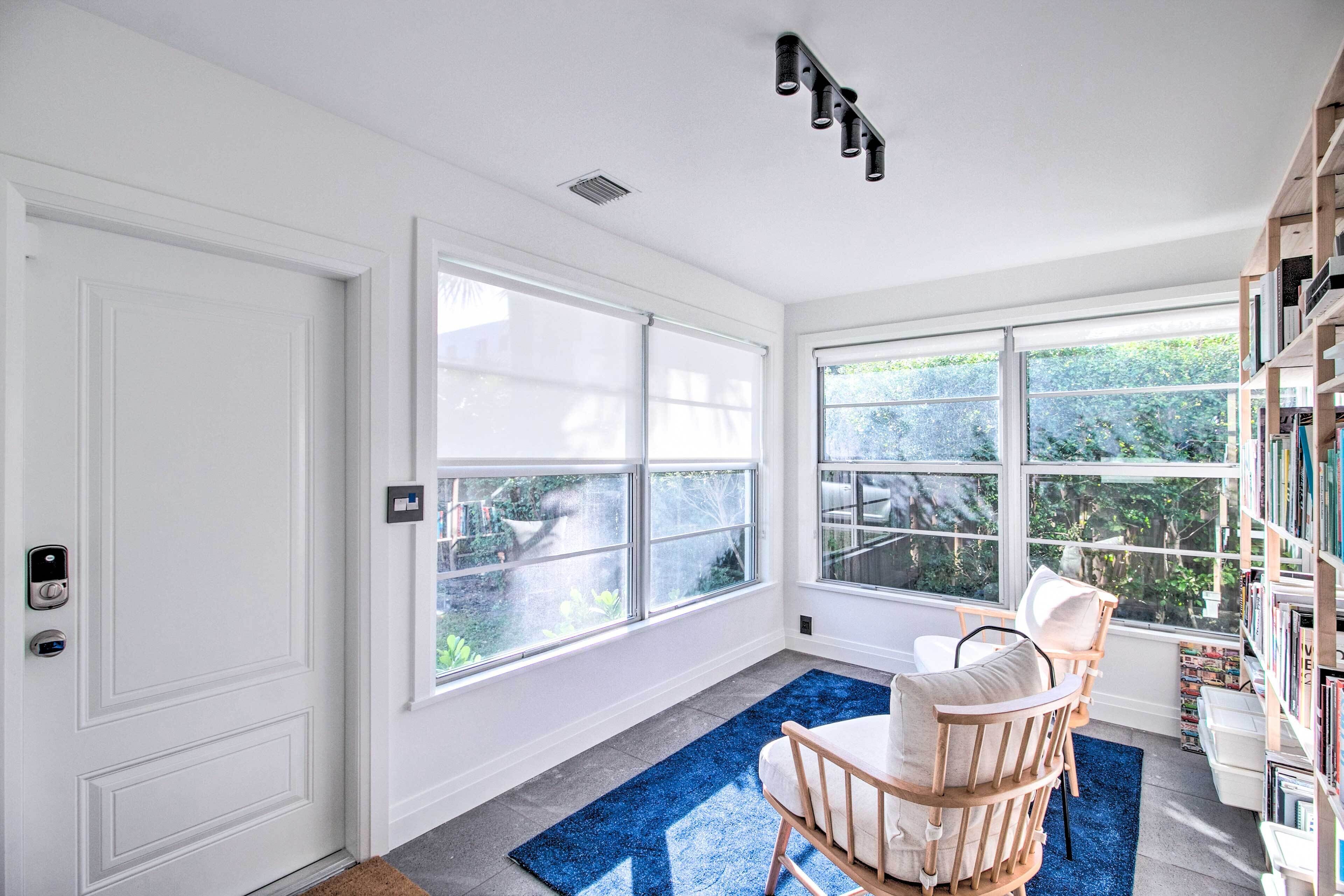 Una habitación luminosa con ventanas amplias, una puerta blanca, un tapete azul, una silla de madera y una estantería.