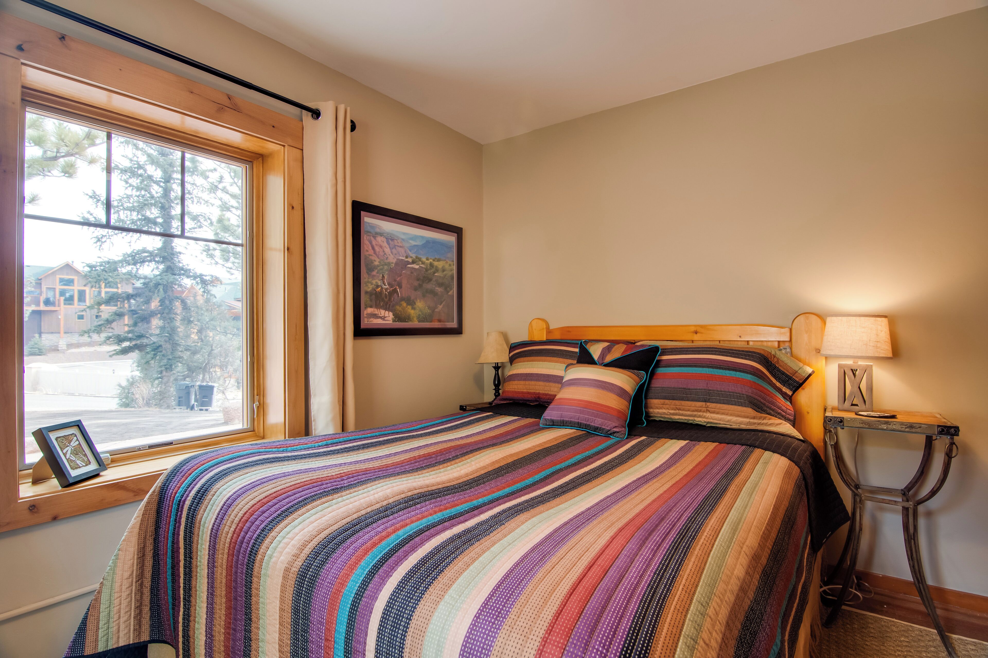 Una cama bien hecha con una colcha a rayas, un cuadro enmarcado en la pared y una ventana con vistas a una casa y árboles.