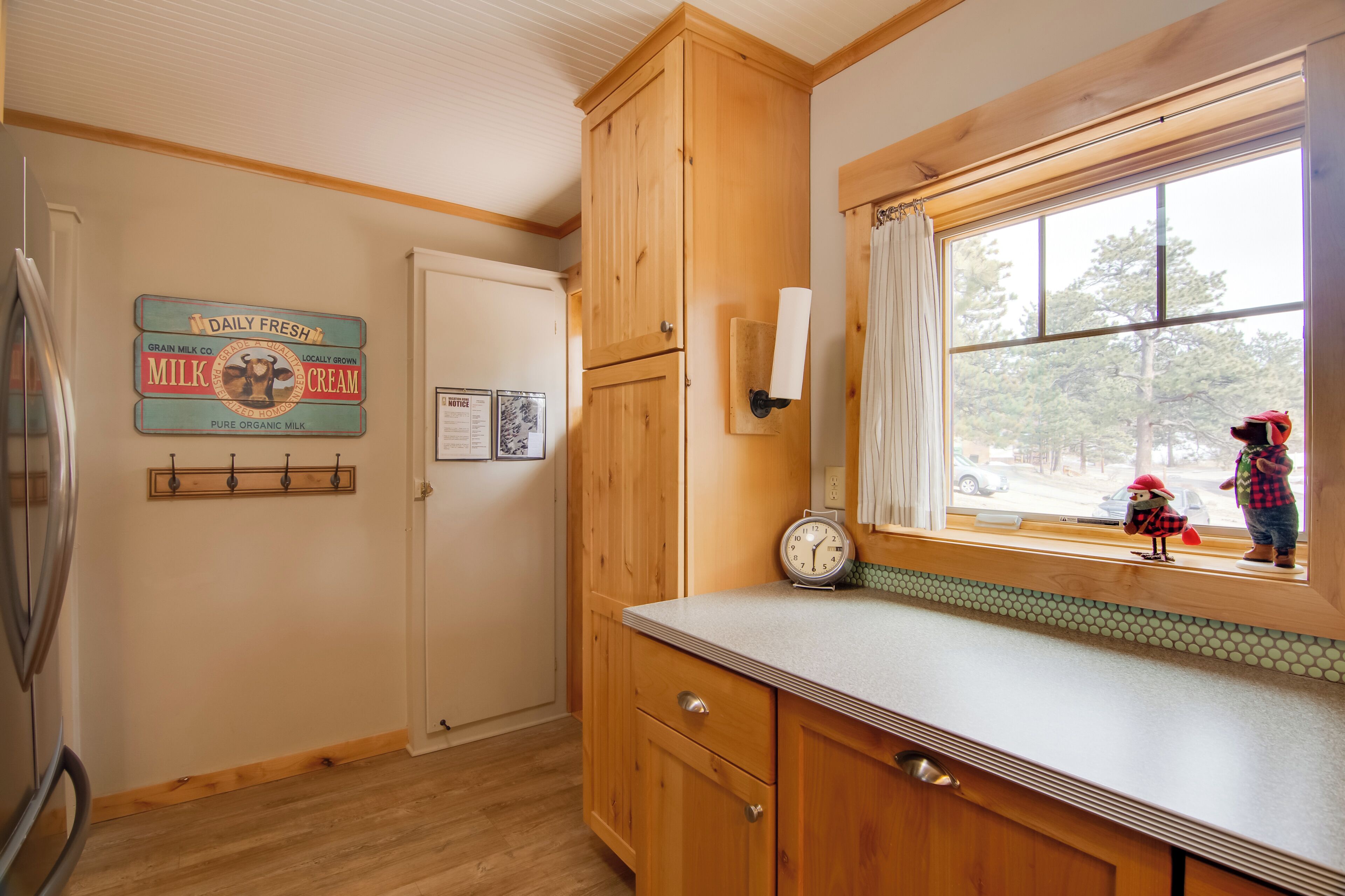 A kitchen with wooden cabinets, a refrigerator, a clock, and a window with a view of the outside.