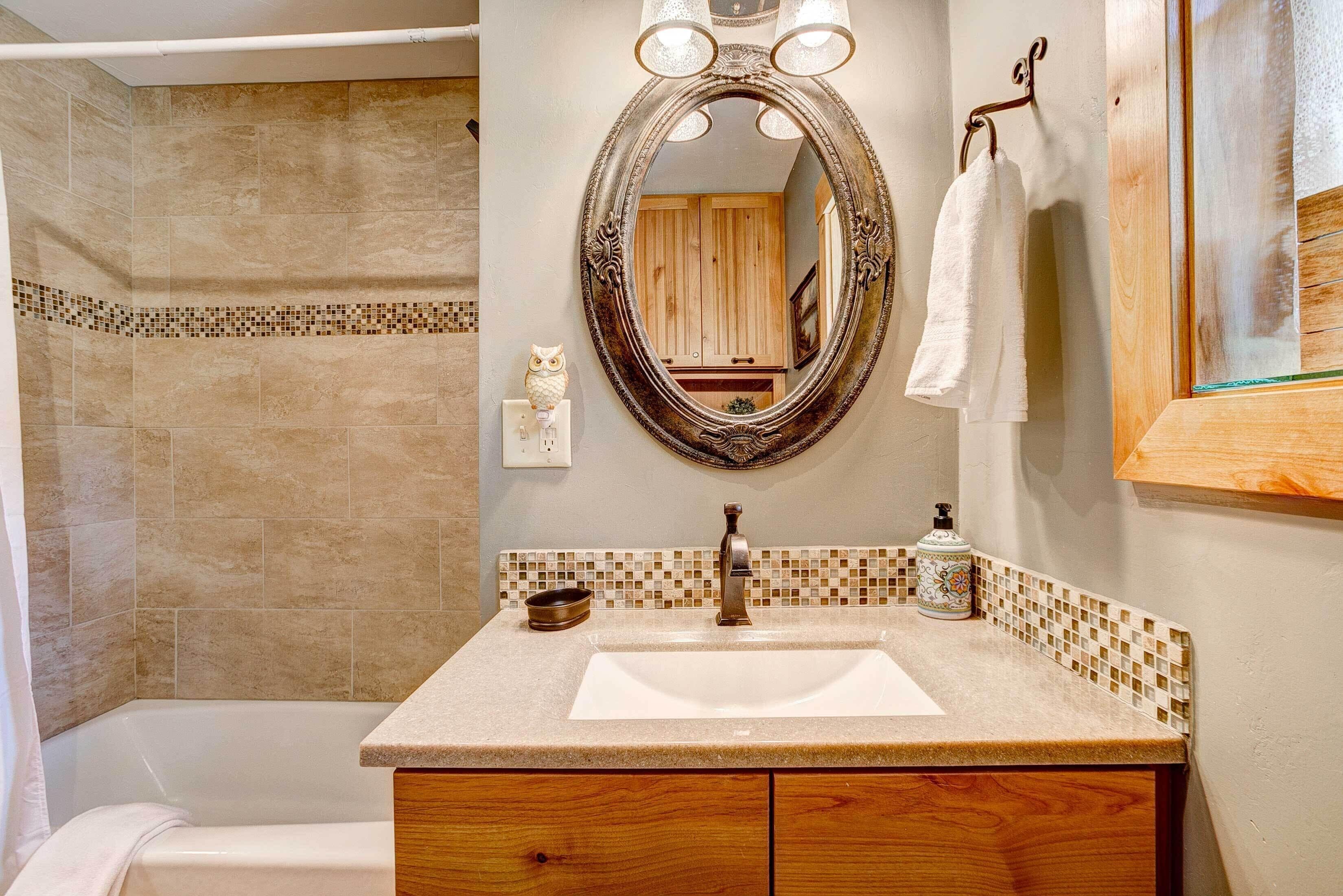 Un baño con pared de azulejos color beige, un lavamanos blanco, un mueble de tocador de madera, un espejo con marco decorativo y una toalla colgada en un perchero.