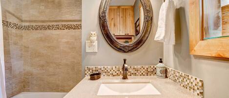 Un baño con pared de azulejos color beige, un lavamanos blanco, un mueble de tocador de madera, un espejo con marco decorativo y una toalla colgada en un perchero.