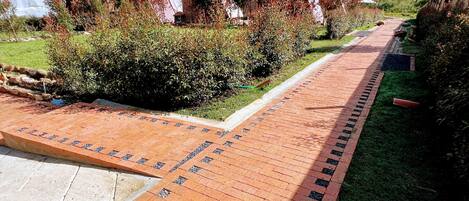 A paved pathway with a brick pattern, bordered by grass and shrubs, leading to a building with a red roof.