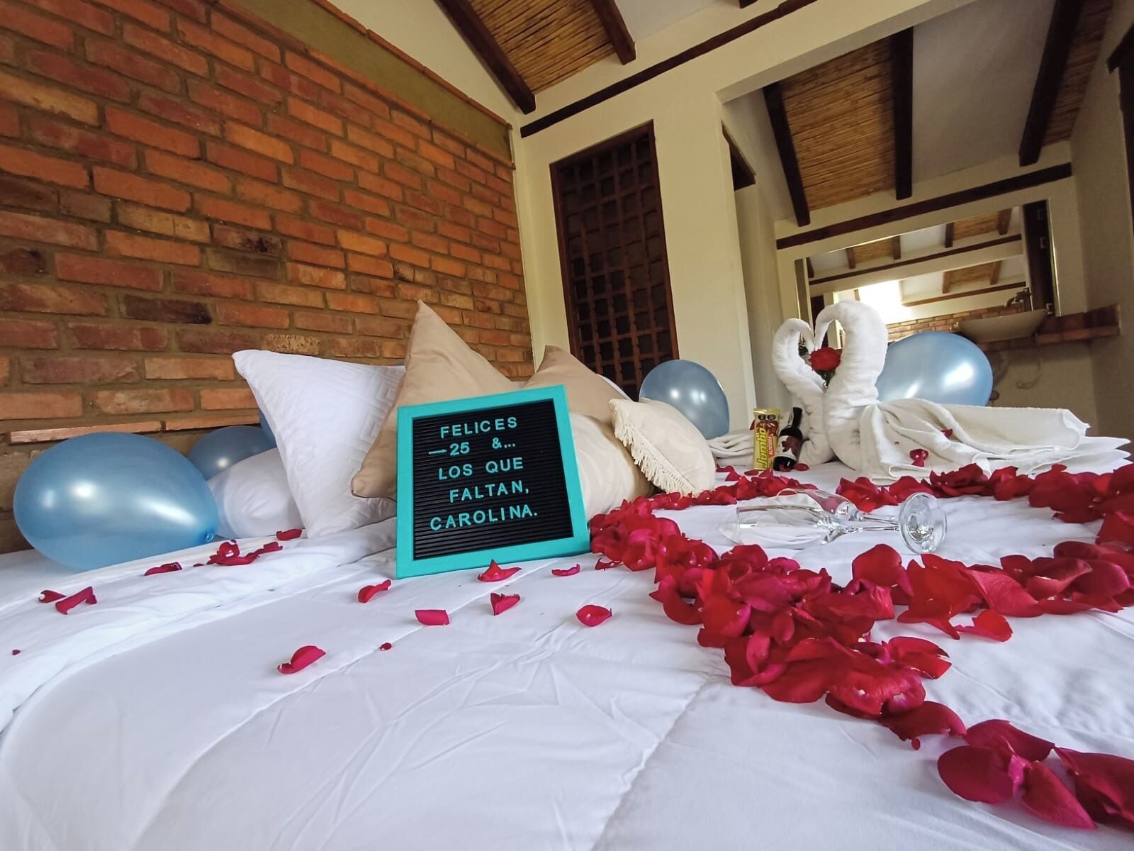 A bed with rose petals, balloons, and a message board in a room with brick walls and wooden beams.
