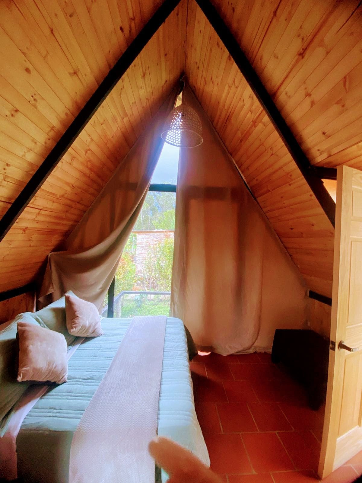 Une chambre mansardée chaleureuse, avec un plafond en pente, un lit avec des oreillers, un luminaire suspendu et une vue par la fenêtre.