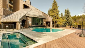 Zona de piscina con terraza de madera, edificio con techo inclinado y bosque al fondo.