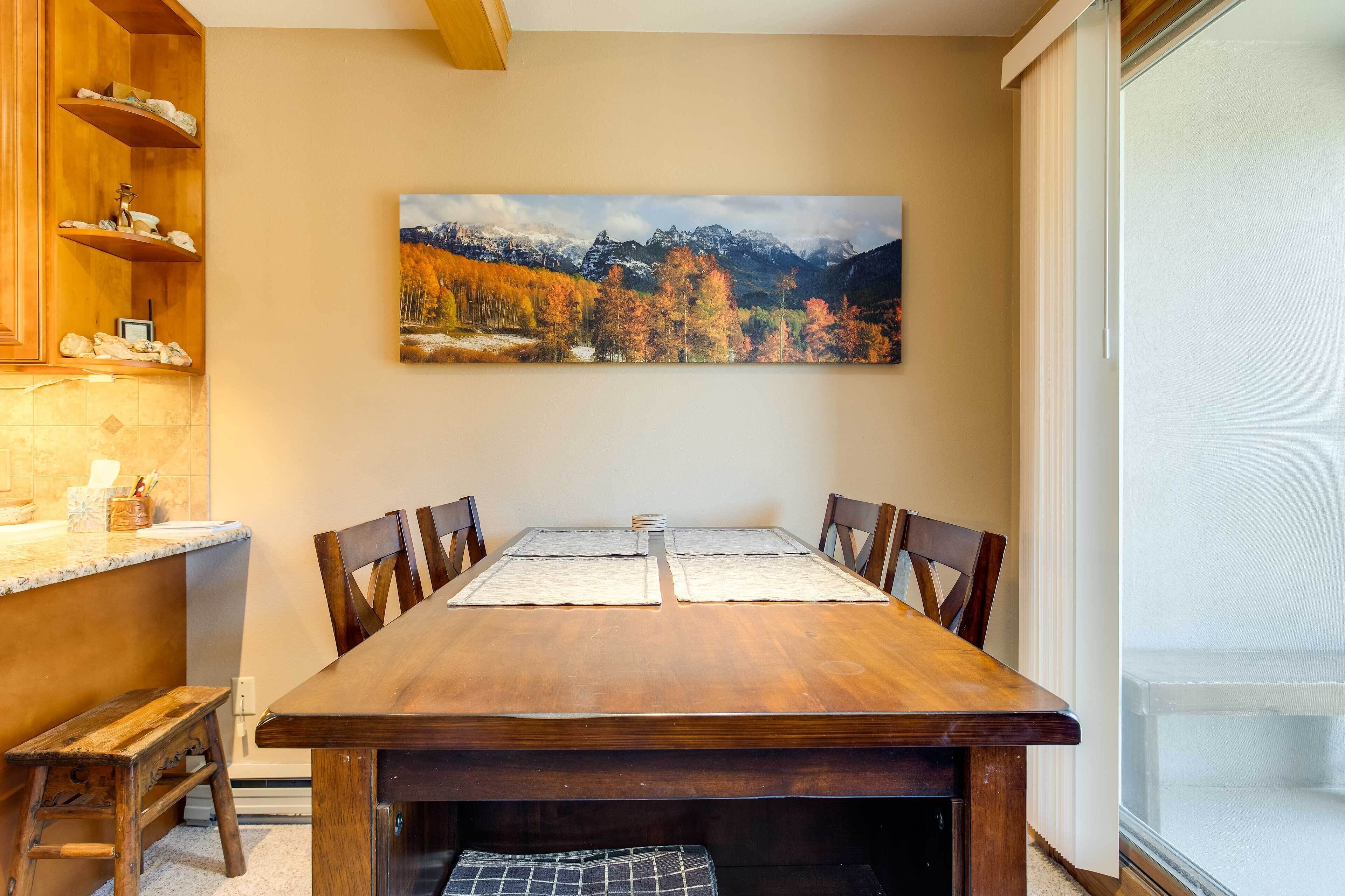 Un comedor con una mesa de madera, sillas y un cuadro de un paisaje montañoso.