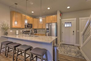 A modern kitchen with wooden cabinets, stainless steel appliances, a marble island with bar stools, pendant lighting, and a view of the living area. - Fraser Retreat, Walk to Ski Shuttle & Main St (Fraser)