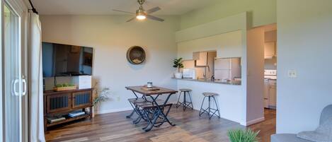 A spacious living area with a wooden floor, a ceiling fan, a dining table with chairs, a kitchen area, and a grey sofa.