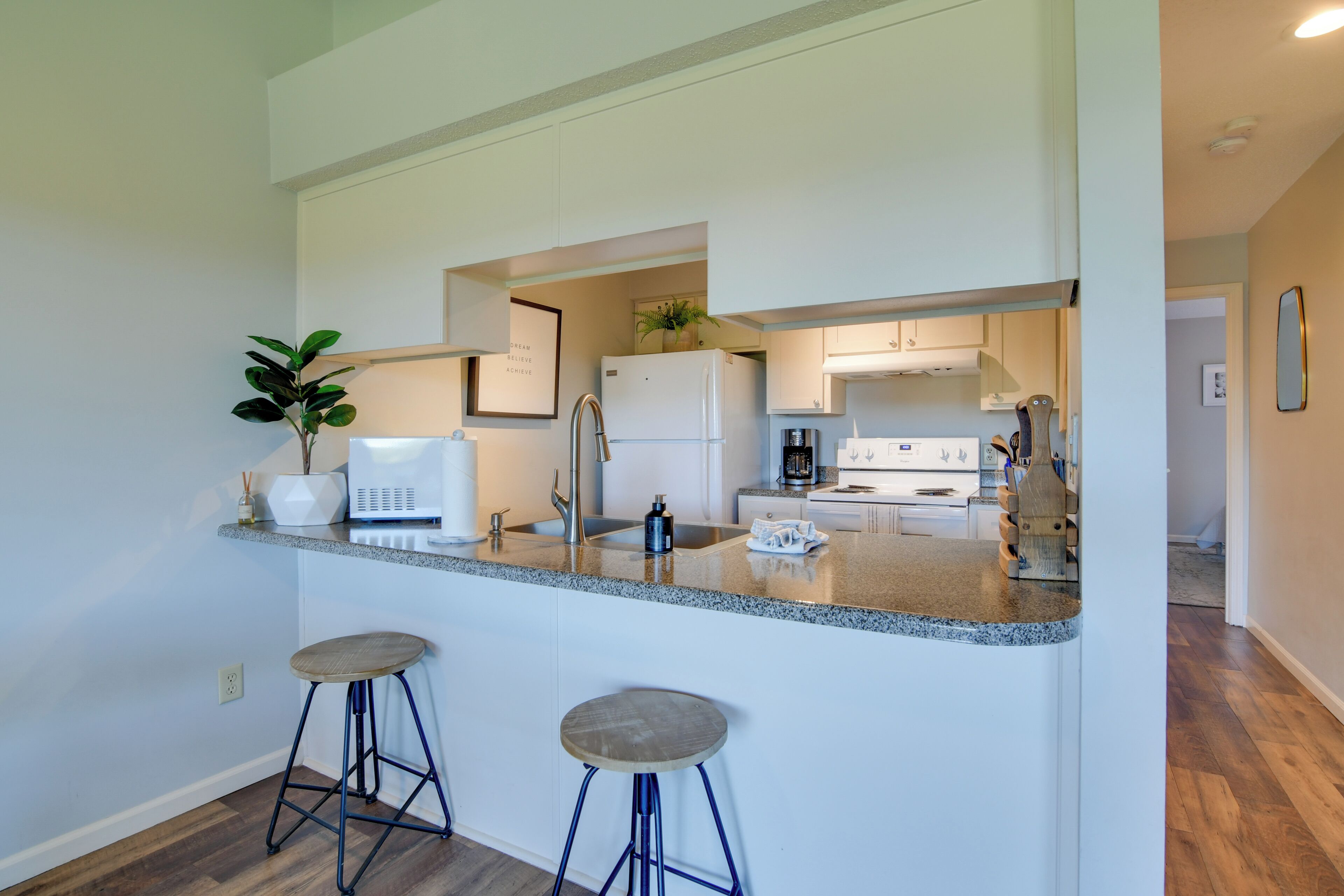 A modern kitchen with white cabinets, granite countertops, and stainless steel appliances. There are two bar stools and a potted plant on the counter.