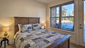 A bedroom with a large window, a wooden bed, a bedside table with a lamp, and a view of a snowy outdoor area.
