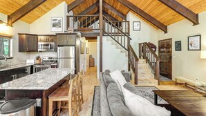 Un vaste espace de vie doté d’un haut plafond et de poutres apparentes, une cuisine équipée d’appareils modernes, un escalier menant à un étage supérieur et un coin salon confortable.