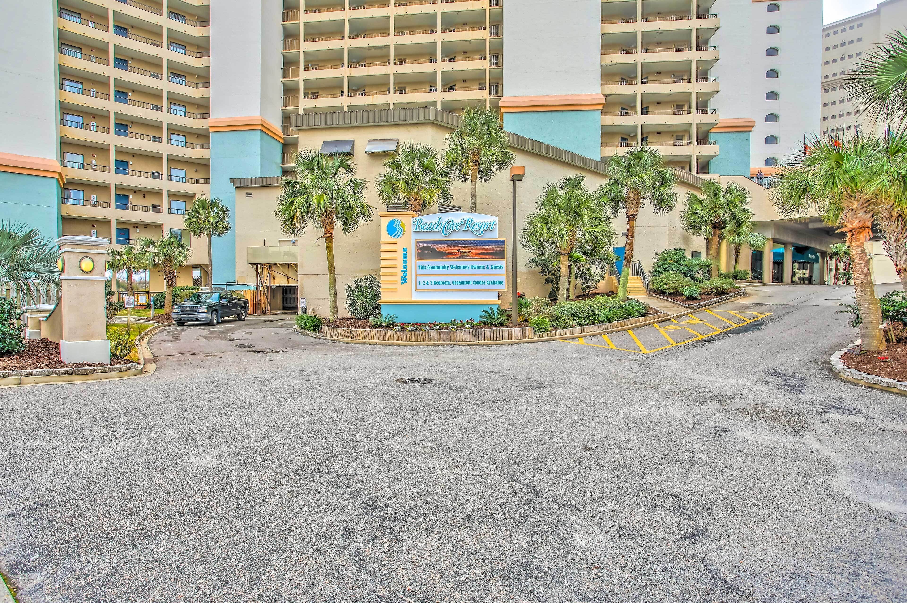 A hotel entrance with a signboard, palm trees, and a parking area.