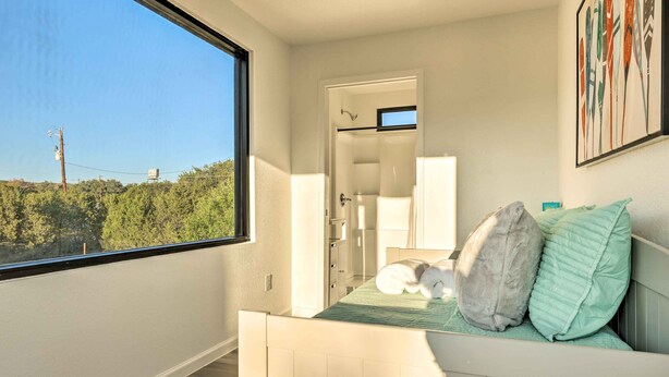 Une chambre avec une grande fenêtre, un lit avec des oreillers, une porte donnant sur une salle de bain et un tableau accroché au mur.