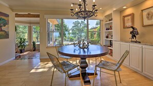 A dining area with a round table, four chairs, a chandelier, and large windows offering a view of greenery. - Luxe Auburn Hideaway w/ Tennis Court & Views! (Auburn)