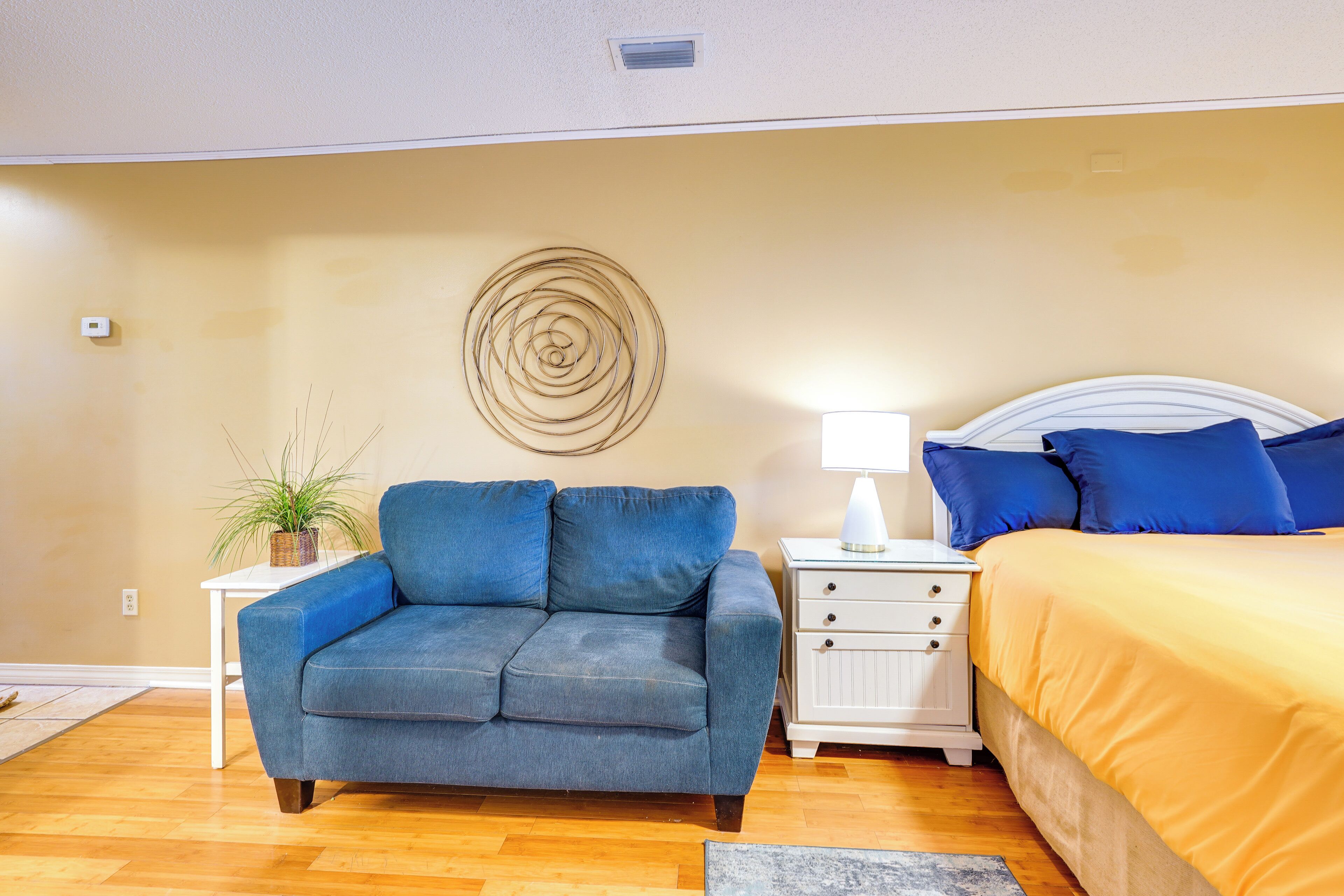Un dormitorio con sofá azul, una mesita de noche blanca con lámpara, una cama con ropa de cama azul y amarilla, y una obra de arte en la pared.