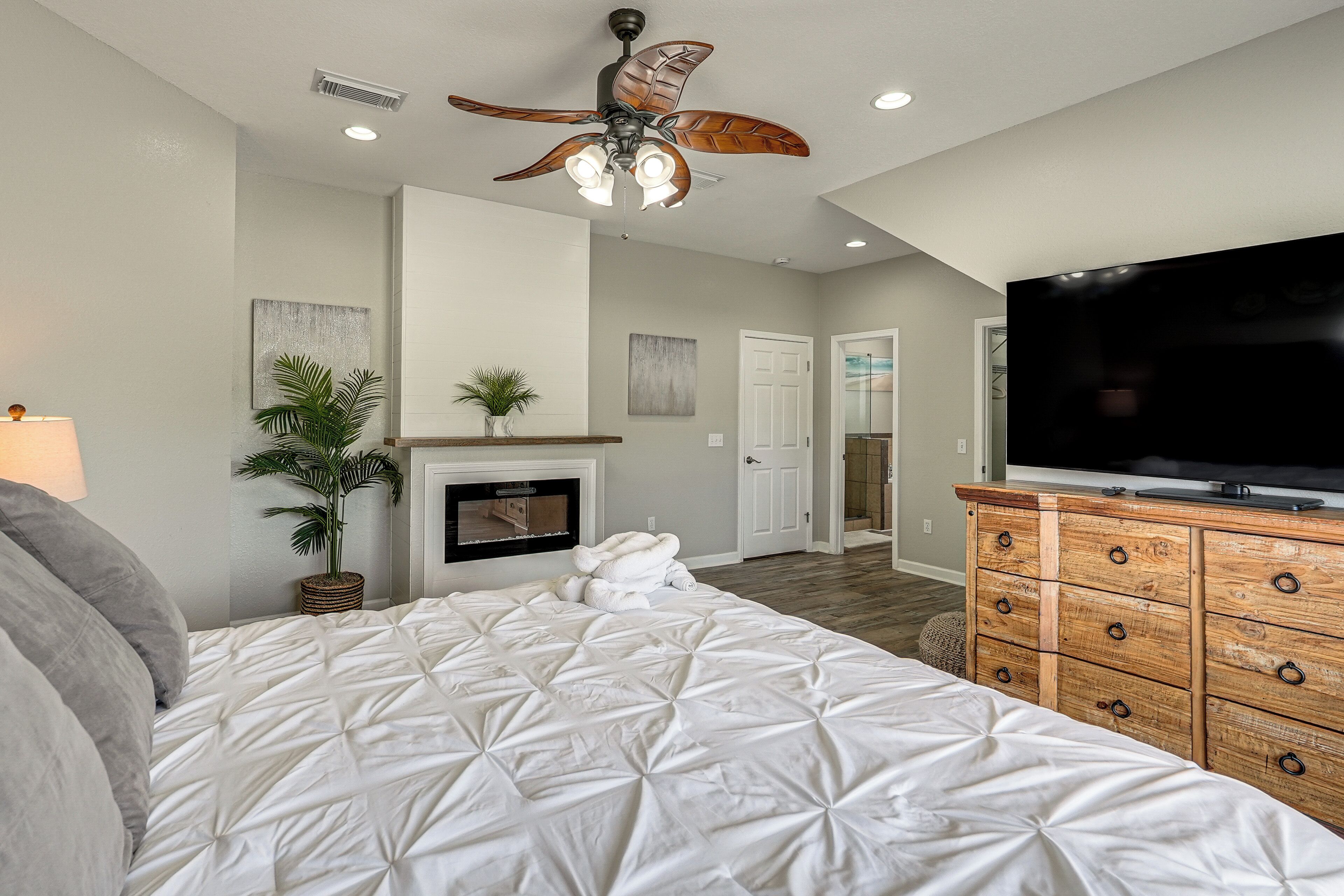 A bedroom with a bed, a ceiling fan, a TV, a dresser, a fireplace, and a door leading to another room.