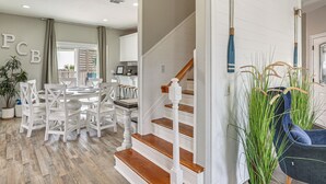 A dining area with a round table and white chairs, a staircase with wooden steps and white railings, and a blue chair with tall green plants. - 'bailey's Beach House Retreat' w/ Game Room! (Panama City Beach)