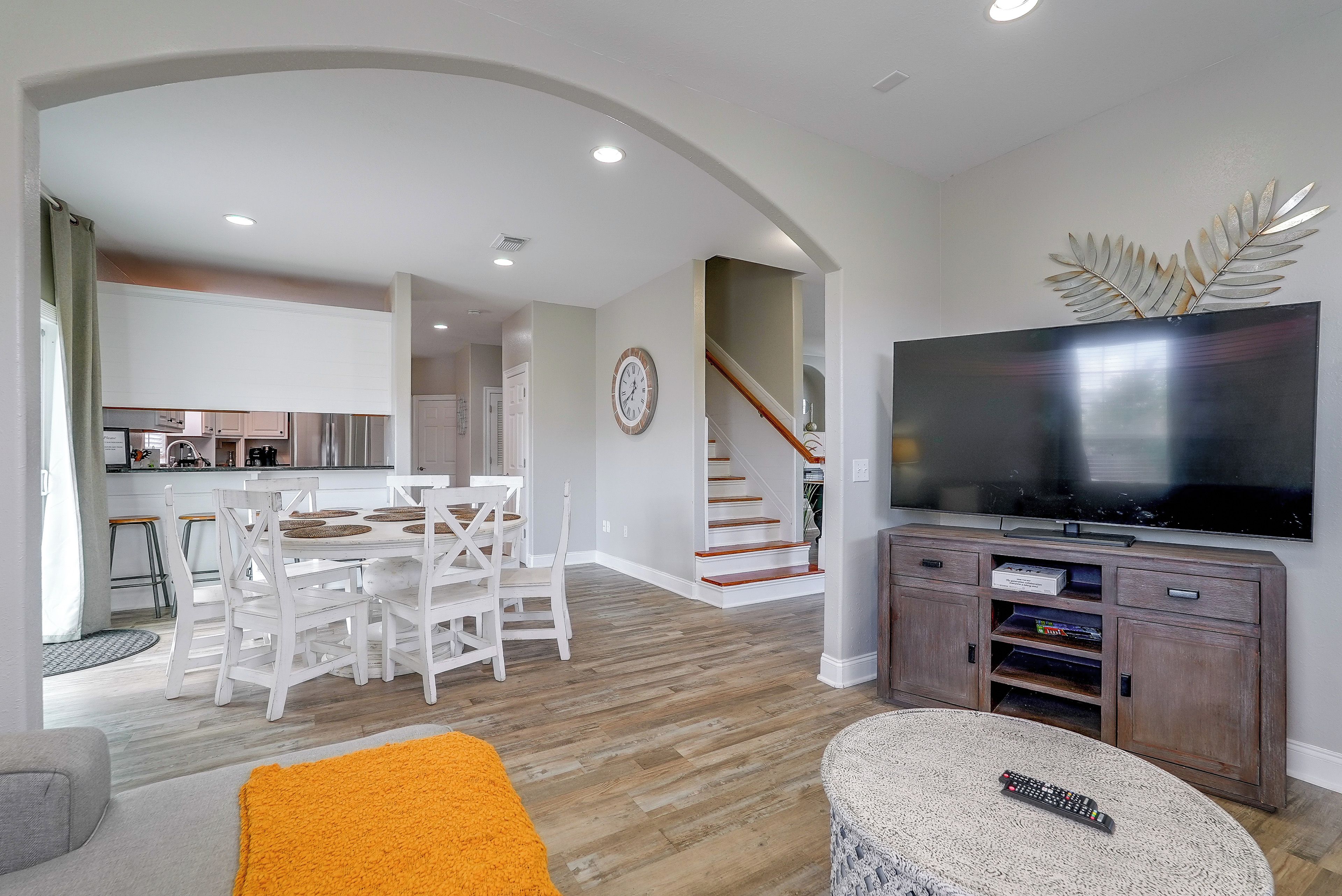 Una sala de estar moderna con televisor, un comedor con mesa y sillas blancas, y una cocina al fondo.