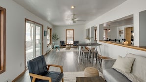 A spacious living area with a dining table, sofa, and chairs. There is a ceiling fan, large windows, and a view of the deck outside.
