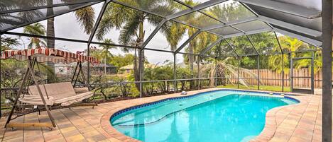 A covered outdoor swimming pool with a swing chair and a view of palm trees.