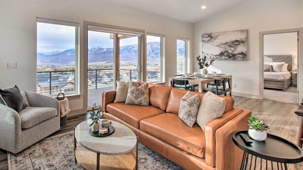 A spacious living room with a large brown leather sofa, a grey armchair, and a round coffee table. There are multiple windows offering a view of mountains and a cityscape. - Eden Home w/ Stunning View ~ 6 Mi to Skiing! (Eden)